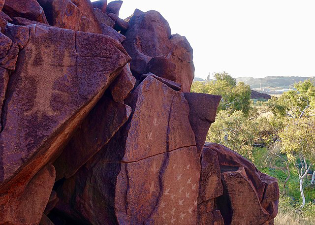 Sur les roches rouges de Murujuga, des gravures ancestrales. L'une d'elle semble avoir une forme humaine. En fond, la forêt aride australienne