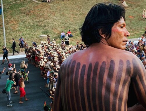 Un individu au premier plan regarde au loin, derrière lui une foule d'autochtones manifeste contre le président jair bolsonaro, à Brasilia au Brésil